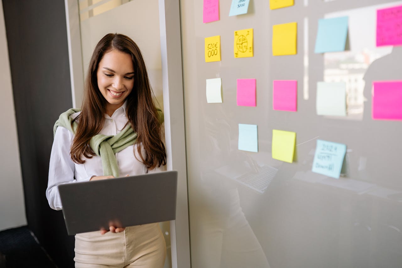 about-01 Smiling woman uses laptop in office, planning with colorful sticky notes on glass wall.